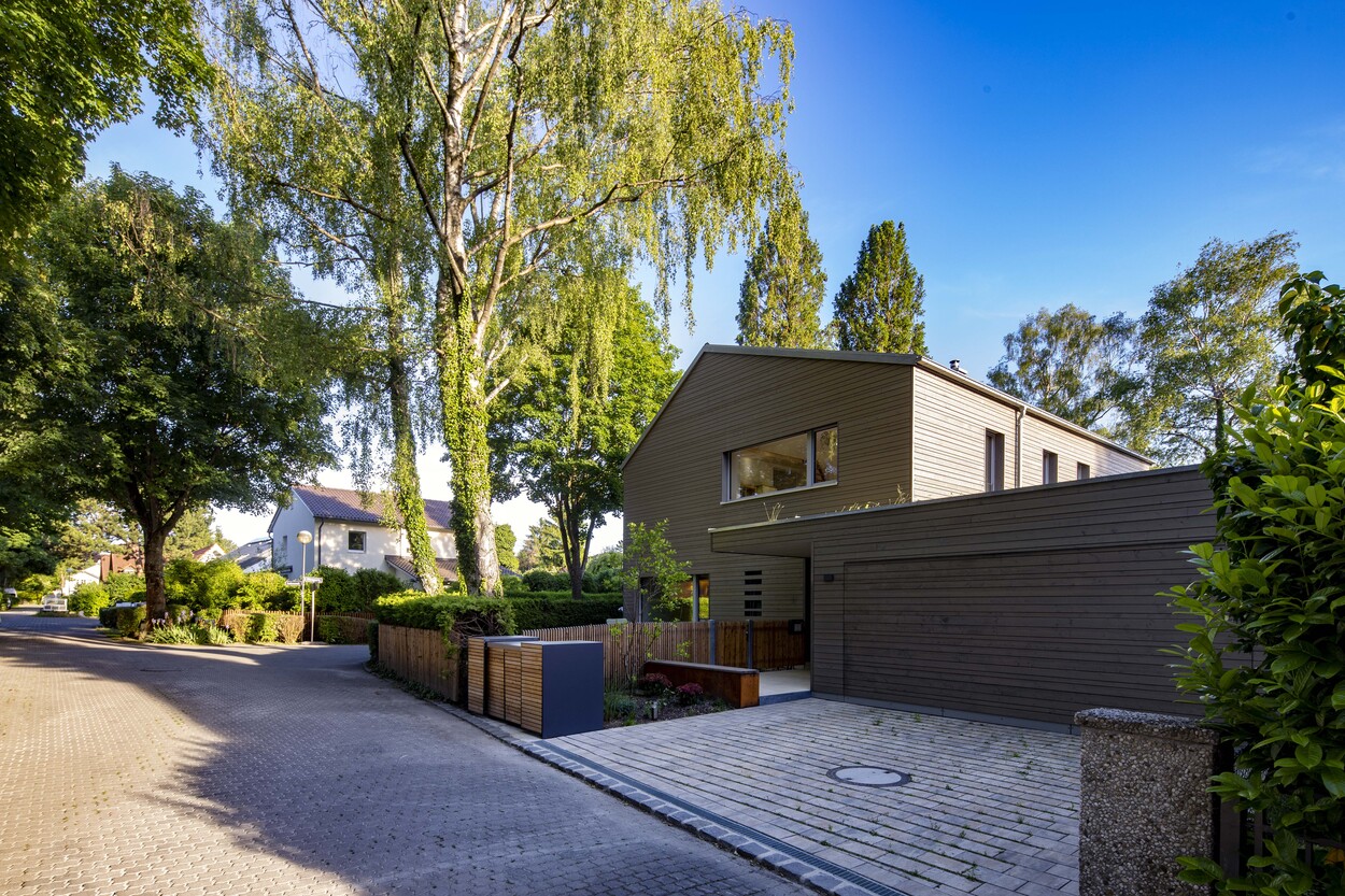 Ein modernes Holzhaus in Gröbenzell steht umgeben von üppigen Bäumen an einer ruhigen, gepflasterten Straße.