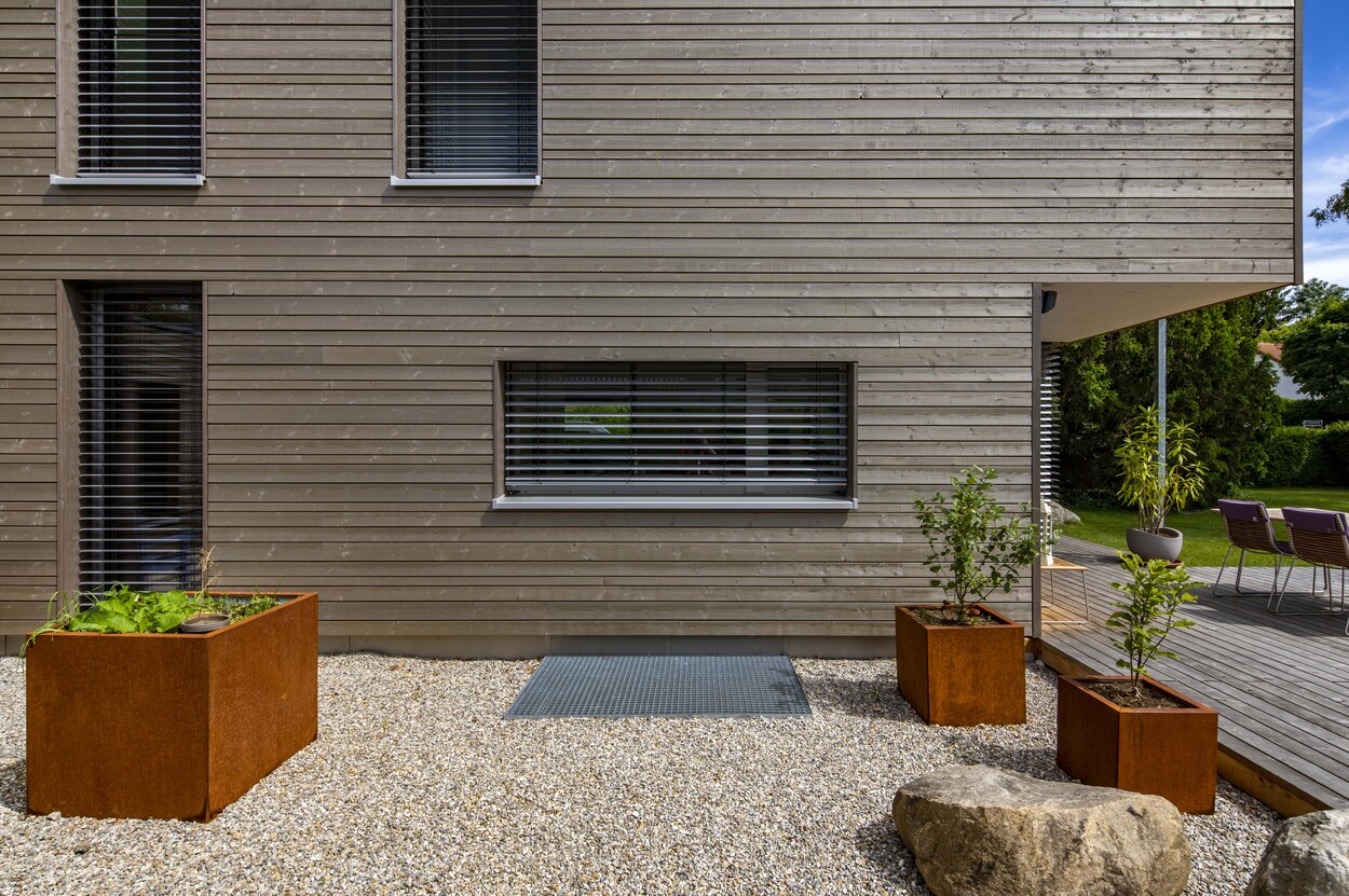 Ein modernes Holzhaus in Gröbenzell mit Kiesweg und rostfarbenen Pflanzkübeln ziert die minimalistische Fassade.