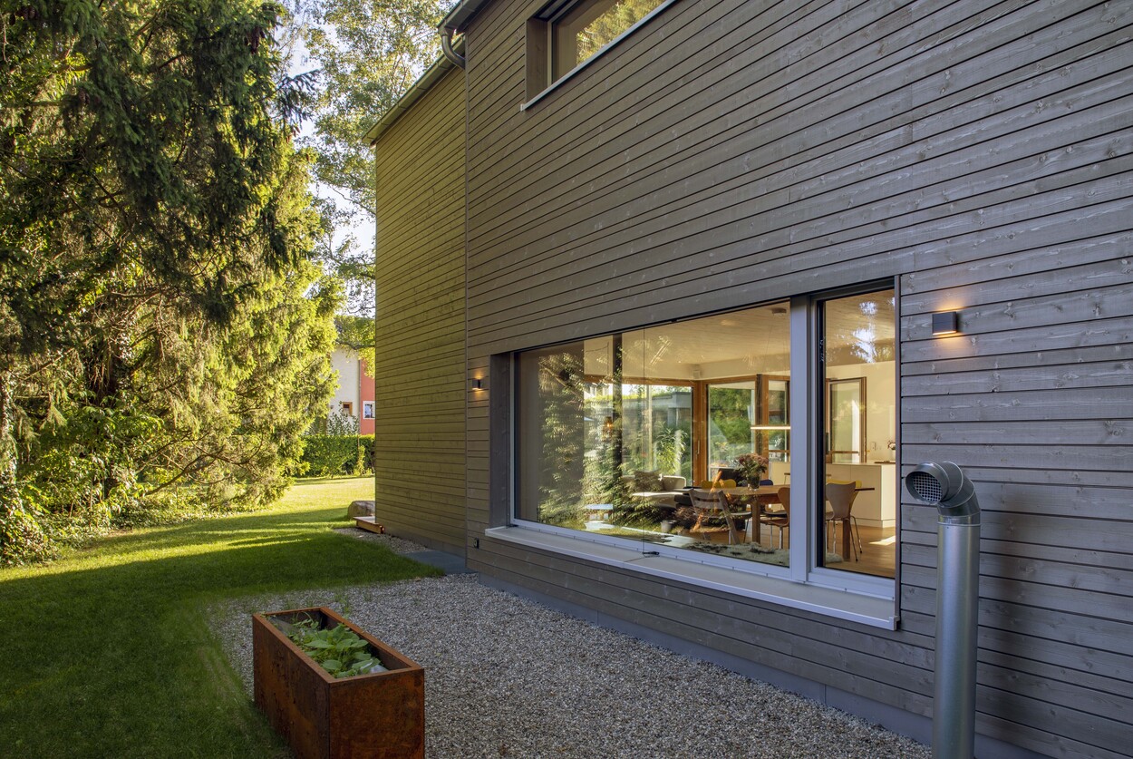 Ein modernes Holzhaus in Gröbenzell zeigt durch große Fenster einen einladenden, hellen Wohnbereich mit grüner Umgebung.