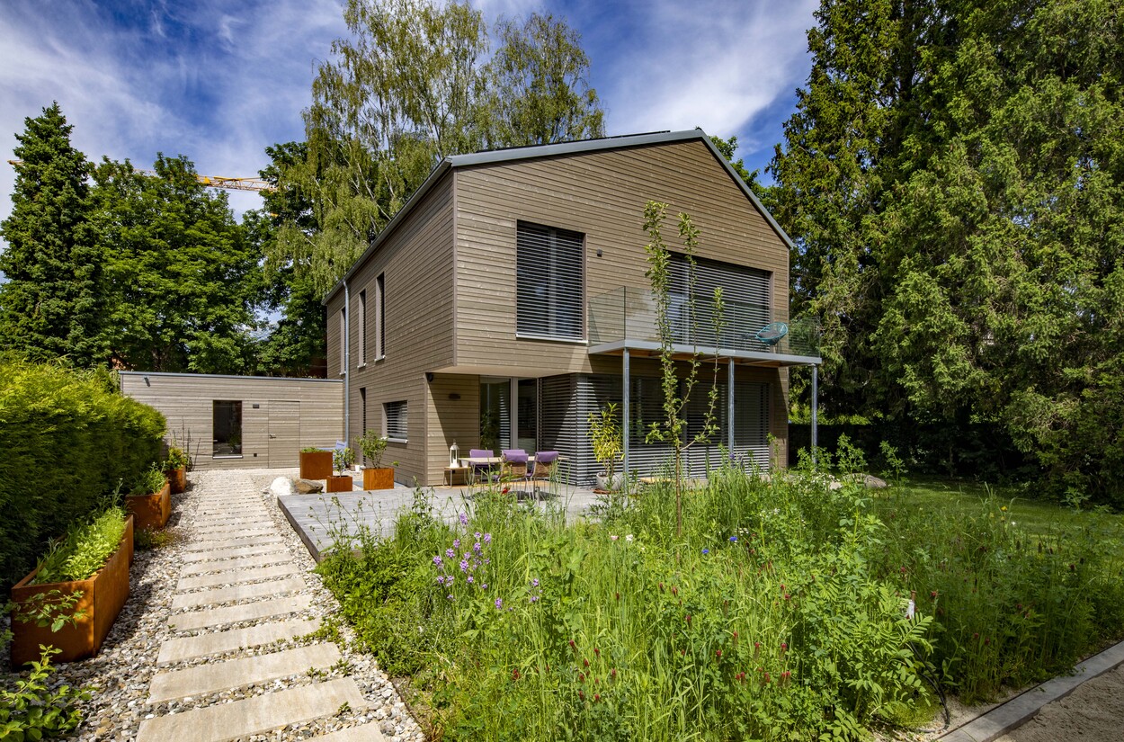 Ein modernes Holzhaus in Gröbenzell ist von einem üppigen Garten und einem gepflasterten Weg umgeben.