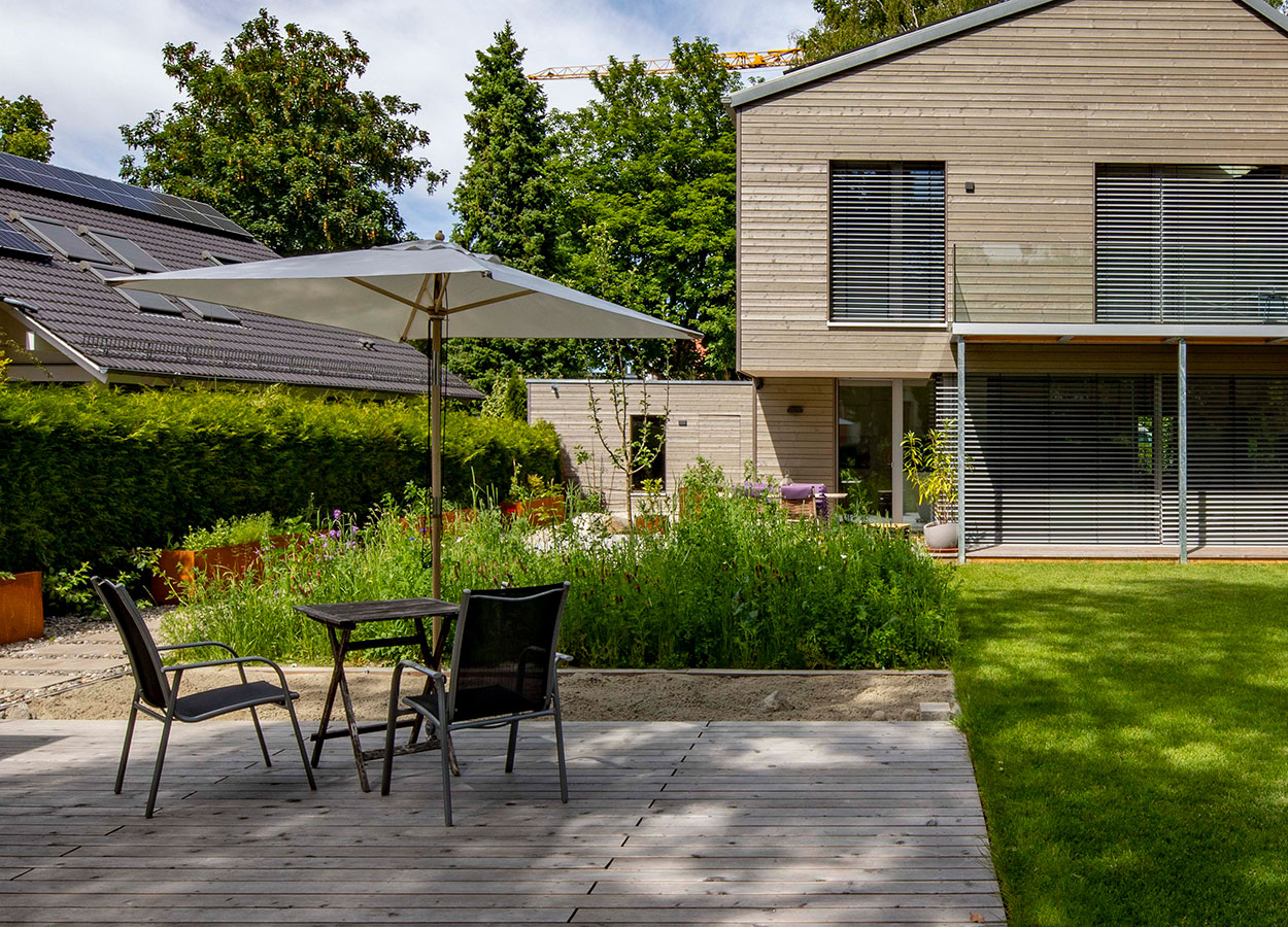 Ein modernes Holzhaus in Gröbenzell mit Terrasse und Gartenmöbeln, umgeben von üppigem Grün und einem Sonnenschirm.