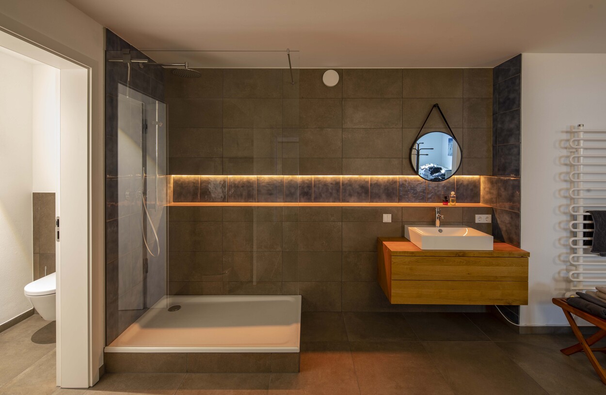 Modernes Badezimmer in einem Holzhaus in Gröbenzell mit beleuchteten Fliesen, einer geräumigen Dusche und stilvollem Waschbecken.