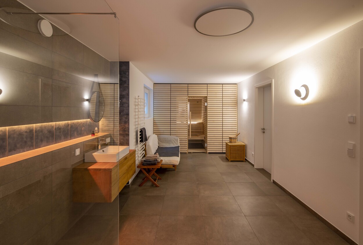Ein modernes Badezimmer in einem Holzhaus in Gröbenzell mit eleganter Beleuchtung und einer einladenden Sauna im Hintergrund.