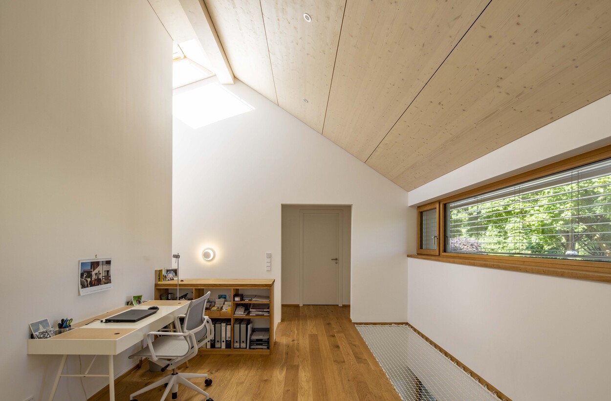 Ein modernes Holzhaus in Gröbenzell zeigt ein lichtdurchflutetes Arbeitszimmer mit weißem Schreibtisch und großem Fenster.