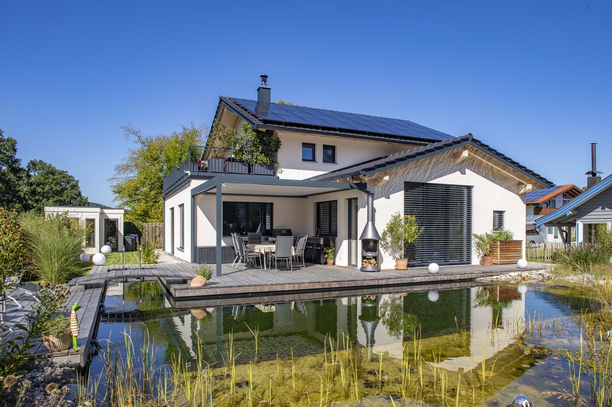 Ein modernes Einfamilienhaus in Prien glänzt mit Solaranlage und Gartenteich unter strahlend blauem Himmel.