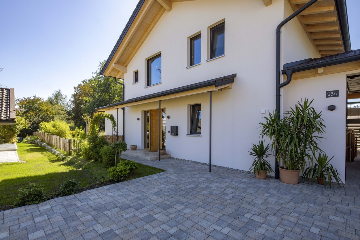 Ein modernes Einfamilienhaus in Prien steht unter strahlend blauem Himmel, umgeben von einem gepflegten Garten und gepflasterter Einfahrt.