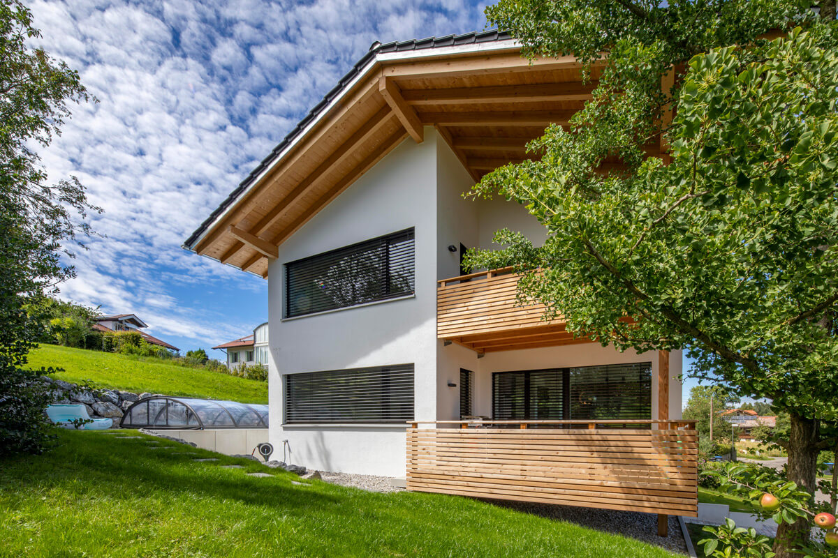 Moderne Holzhaus-Außenansicht in Rimsting mit Balkon, umgeben von grünem Garten und blauem Himmel.