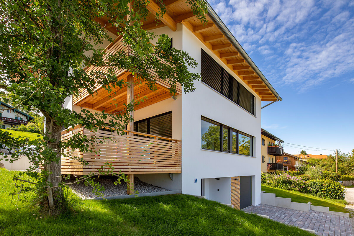 Ein modernes Holzhaus in Rimsting mit großzügiger Holzveranda steht in grüner Umgebung unter klarem Himmel.