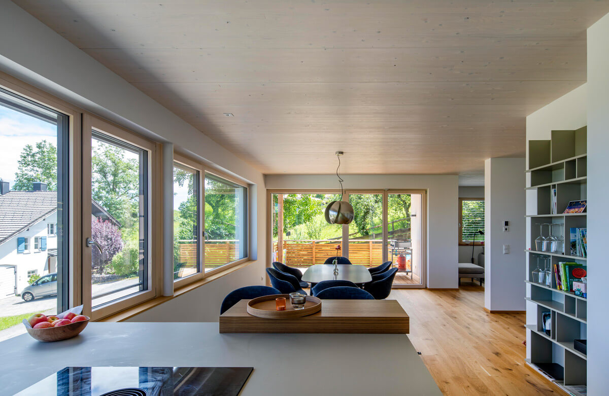 Ein modernes Esszimmer in einem Holzhaus in Rimsting wird von großen Fenstern mit viel Licht auf den Esstisch erhellt.
