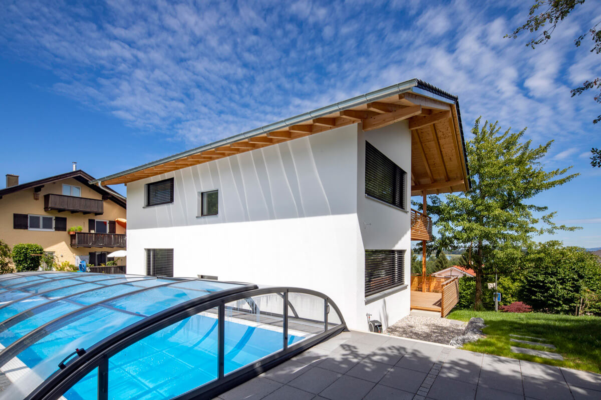 Ein modernes Holzhaus in Rimsting steht neben einem überdachten Pool, umgeben von grünen Bäumen und blauem Himmel.