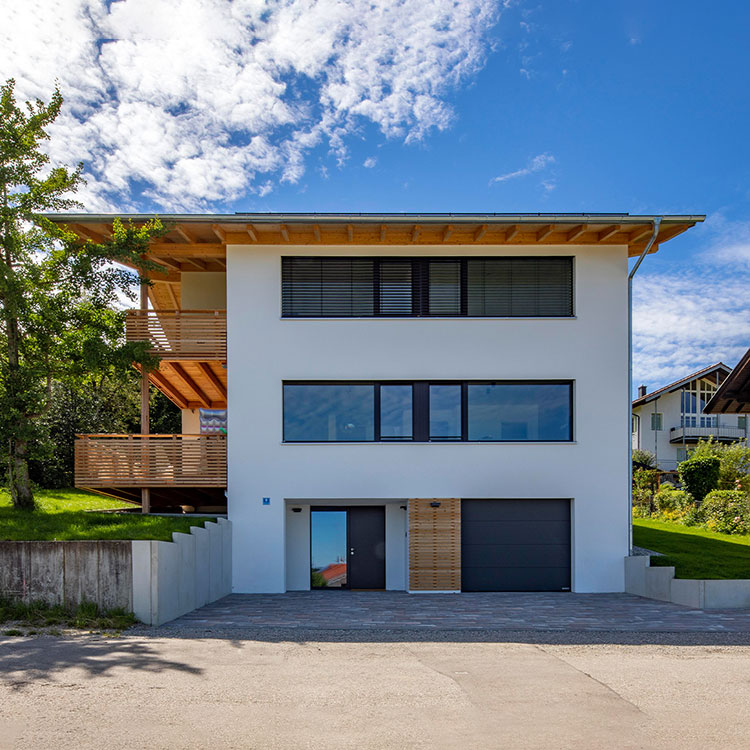 Ein modernes Holzhaus in Rimsting mit weißer Fassade und großen Fenstern steht unter einem strahlend blauen Himmel.