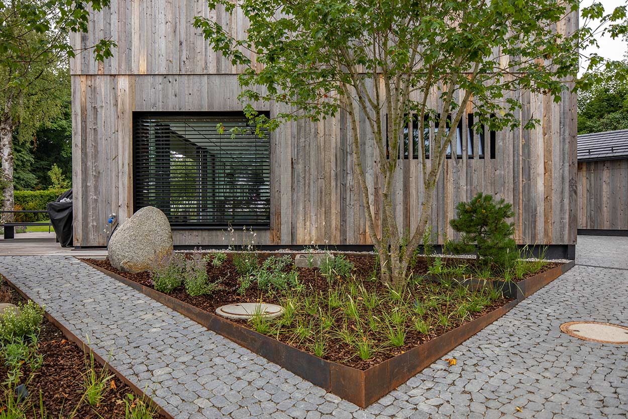 Ein modernes Holzhaus mit großen Fenstern steht neben einem gepflegten Garten mit gepflastertem Gehweg und Bäumen.