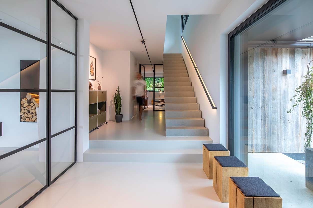 Ein moderner, lichtdurchfluteter Flur mit Treppe und Holzhockern führt zu einem Raum mit großen Glasfenstern.