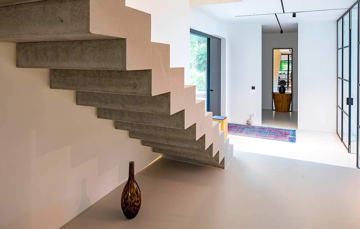 Eine moderne Betontreppe führt in einem hellen Raum mit minimalistischem Stil und buntem Teppich nach oben.