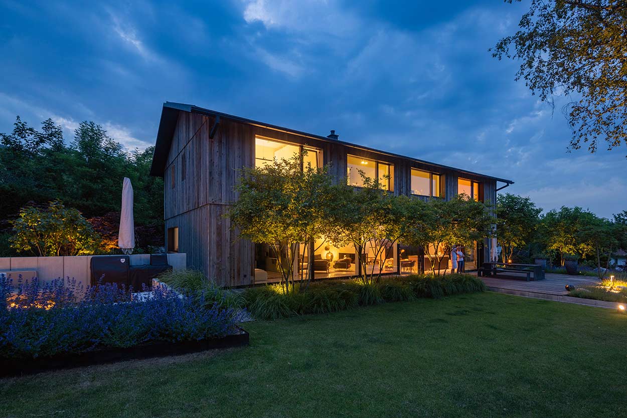Ein modernes Holzhaus leuchtet abends warm im gepflegten Garten, umgeben von blühenden Pflanzen und Bäumen.