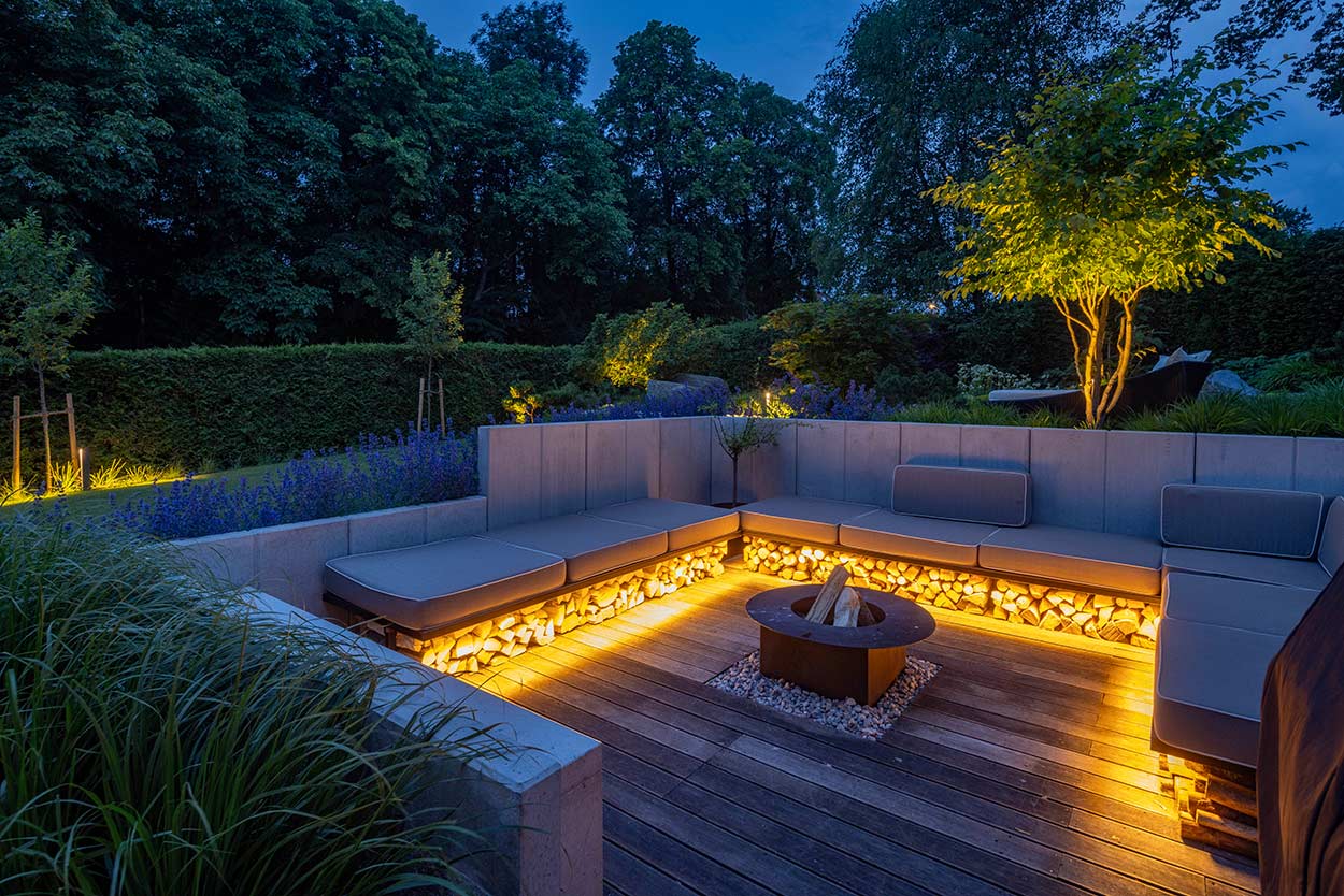 Ein moderner Garten mit beleuchteter Sitzgruppe und Feuerstelle lädt in der Abenddämmerung zum Entspannen ein.