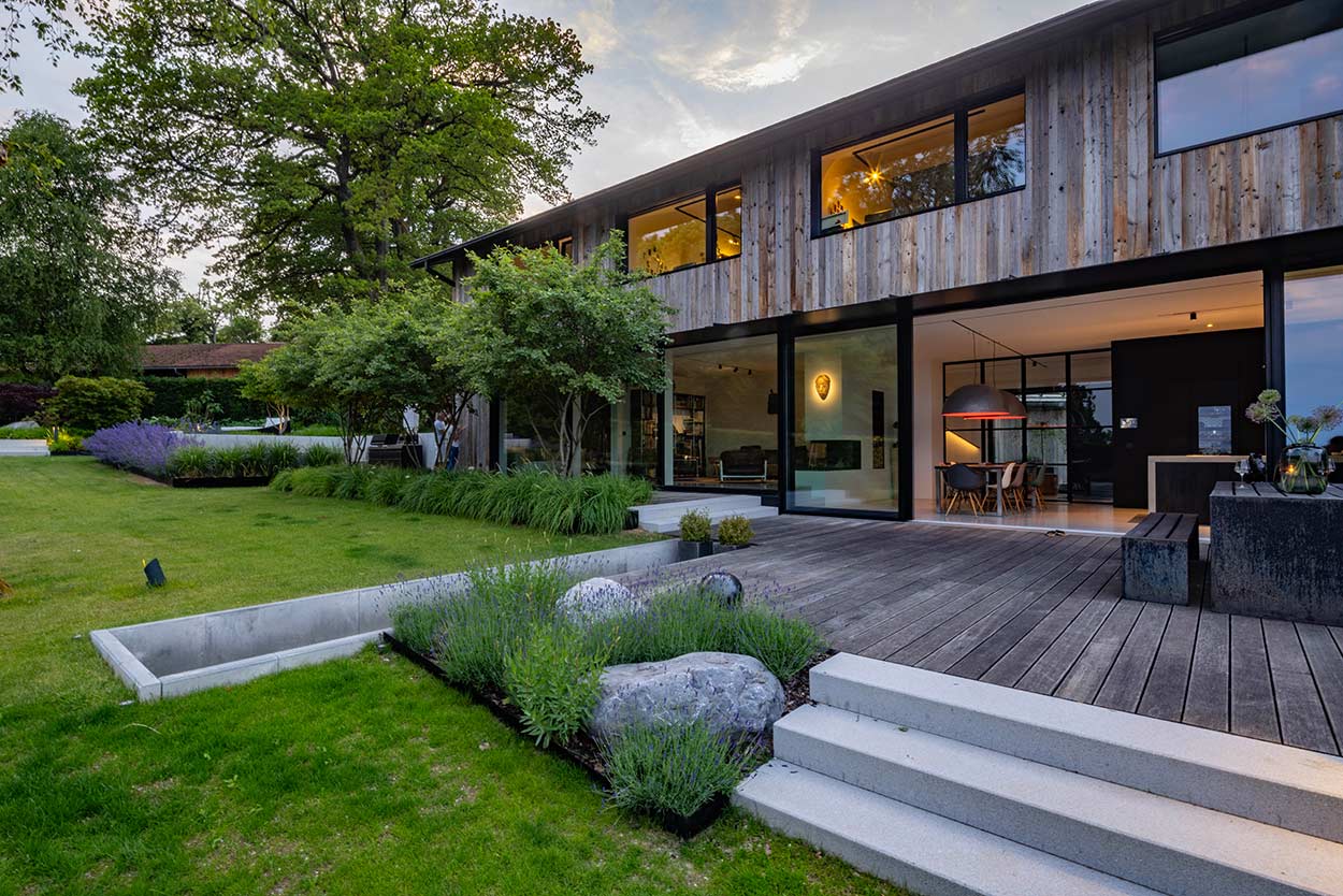 Ein modernes Haus mit Holzfassade und großen Fenstern steht in einem gepflegten Garten mit üppiger Vegetation.