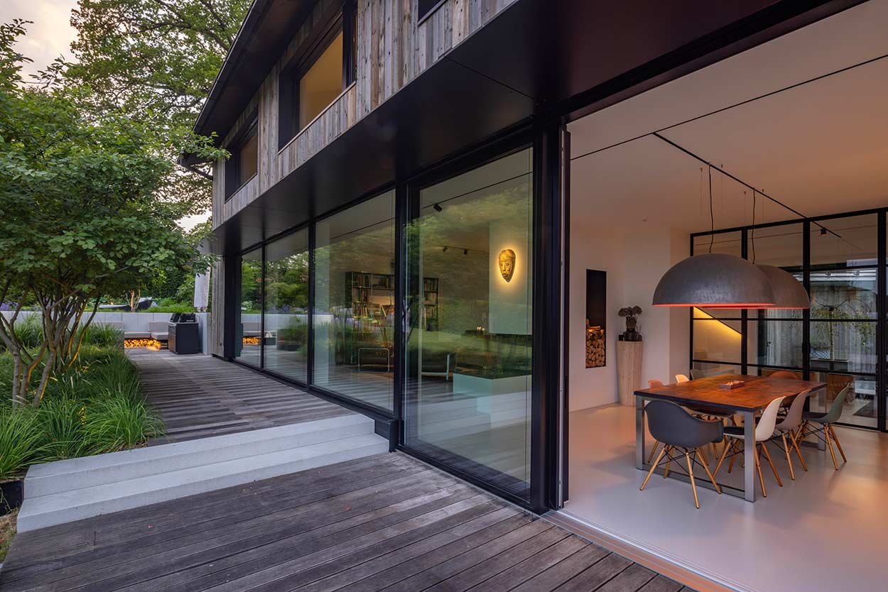 Moderne Villa mit großer Glasfront, einladendem Essbereich und Holzterrasse, umgeben von üppiger Vegetation.