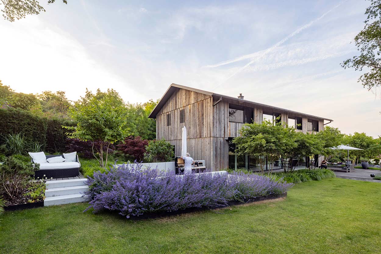 Ein modernes Holzhaus erhebt sich in einem grünen Garten, umgeben von Lavendelbeeten unter einem klaren Himmel.