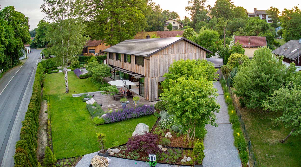Ein modernes Holzhaus mit üppigem Garten und Terrasse liegt idyllisch an einer ruhigen Straße in grüner Umgebung.