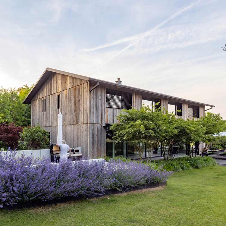 Ein modernes Holzhaus thront in einem gepflegten Garten mit blühenden Lavendelsträuchern und einem einladenden Grillbereich.