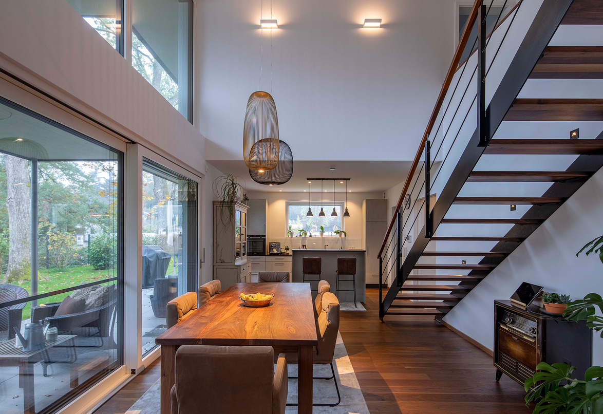 Ein modernes Esszimmer im Energiesparhaus nahe München zeigt eine Holztreppe und große Fenster mit Blick auf den Garten.