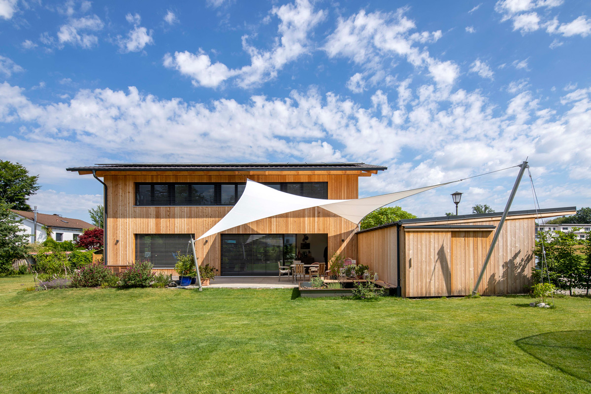 Ein modernes Holzhaus in Neubeuern mit Sonnensegel steht in einem gepflegten Garten unter strahlend blauem Himmel.