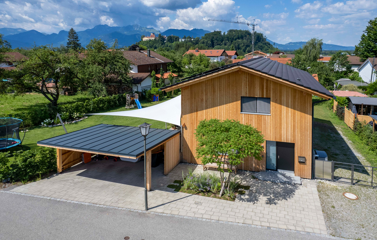 Ein modernes Holzhaus mit Carport in Neubeuern steht vor einer malerischen Bergkulisse an einem sonnigen Tag.