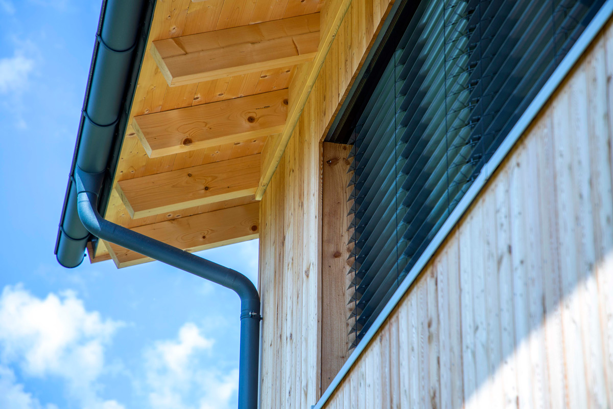 Detailansicht eines Holzhauses in Neubeuern zeigt modernes Fensterdesign und Dachrinne unter klarem Himmel.