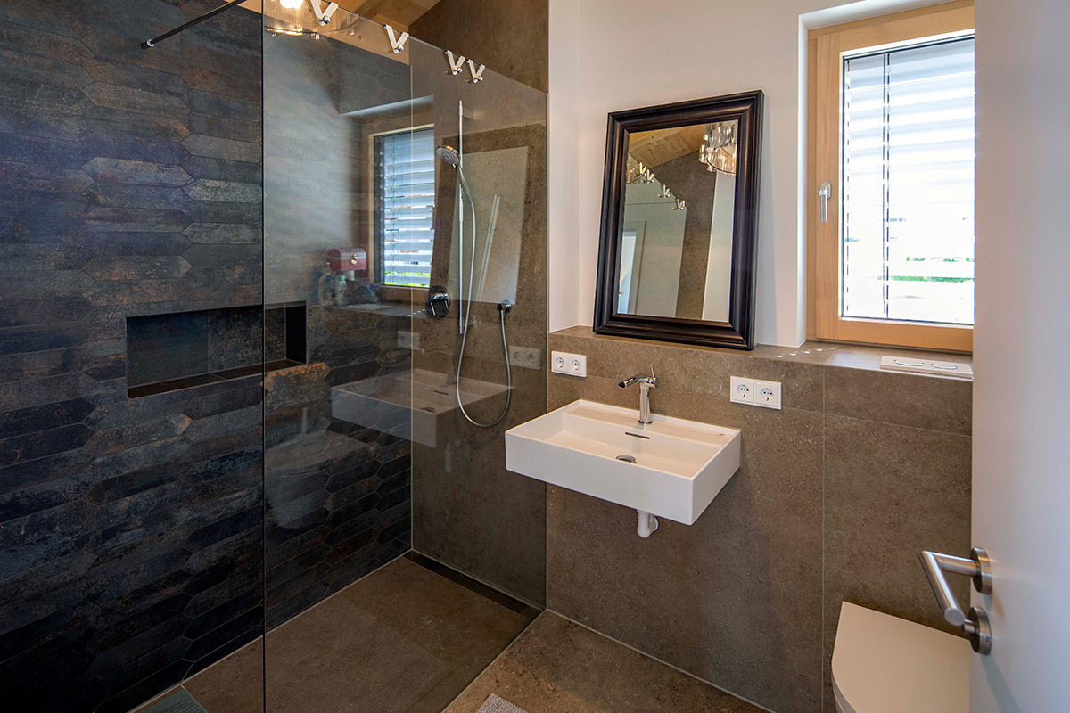 Modernes Badezimmer mit großer Glasdusche, weißem Waschbecken und Fenster mit Holzrahmen in einem Holzhaus in Neubeuern.