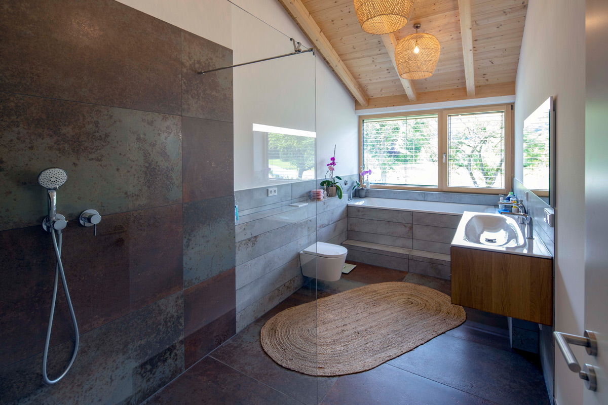 Ein modernes Bad in einem Holzhaus in Neubeuern mit Dusche und Badewanne, durchflutet von Tageslicht durch ein großes Fenster.