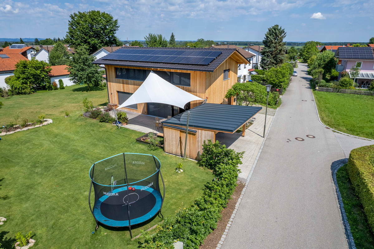 Ein modernes Holzhaus mit Solarzellen steht in einem großen Garten in Neubeuern, neben einer Straße mit Trampolin.