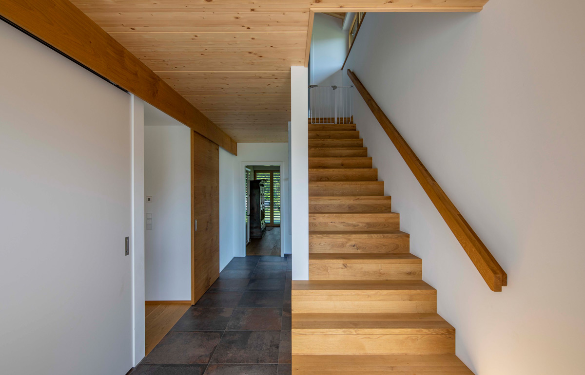 Eine elegante Holztreppe führt in einem modernen Holzhaus in Neubeuern nach oben, flankiert von minimalistischen Wänden.