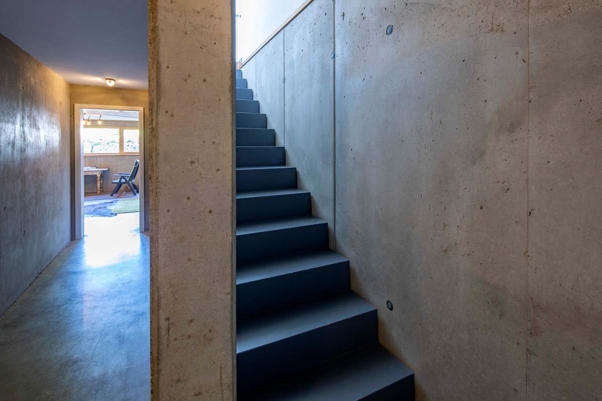 Eine moderne Betontreppe führt in einem Holzhaus in Neubeuern zu einem hellen Raum mit großen Fenstern.
