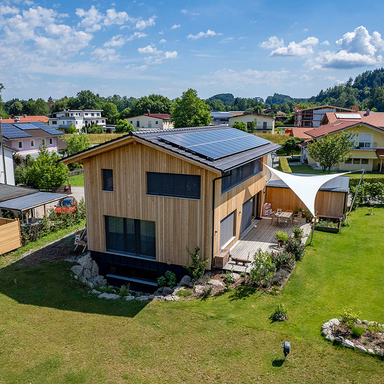 Ein modernes Holzhaus in Neubeuern mit Solarmodulen auf dem Dach steht idyllisch in einer grünen, sonnigen Umgebung.