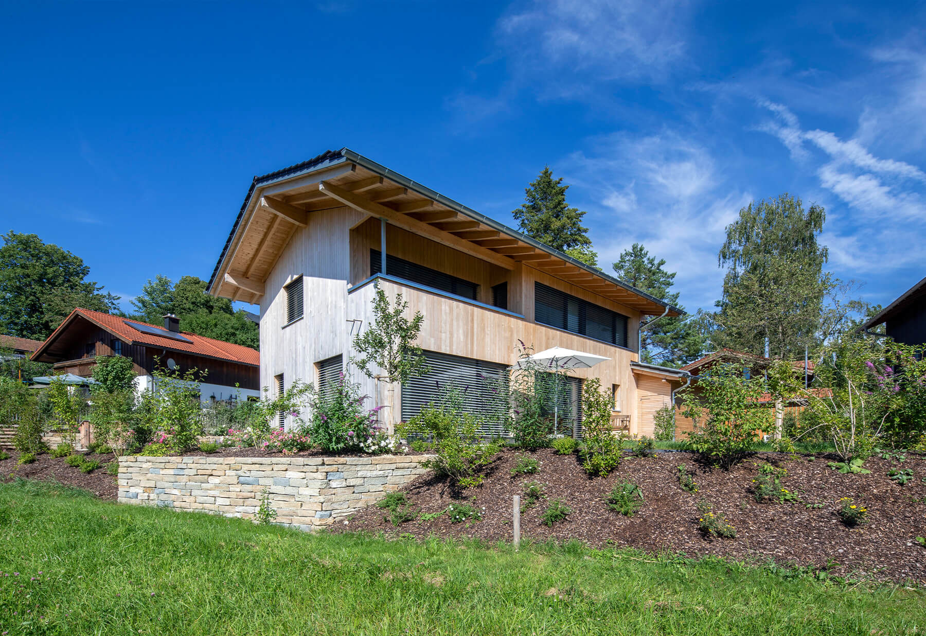 Ein modernes Holzhaus in Chieming steht unter blauem Himmel, umgeben von üppigem Garten und gepflegtem Rasen.
