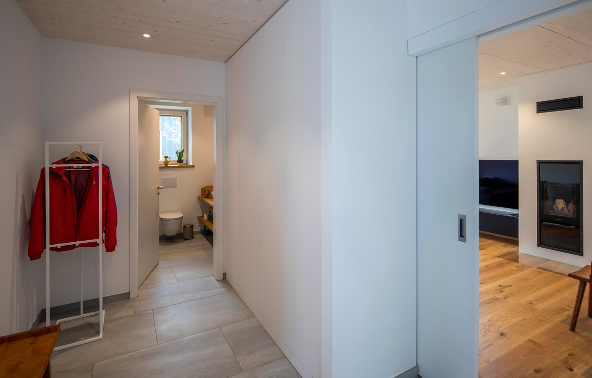 Ein heller Flur im Holzhaus in Chieming zeigt eine rote Jacke an der Garderobe und führt zu einem modernen Wohnzimmer.