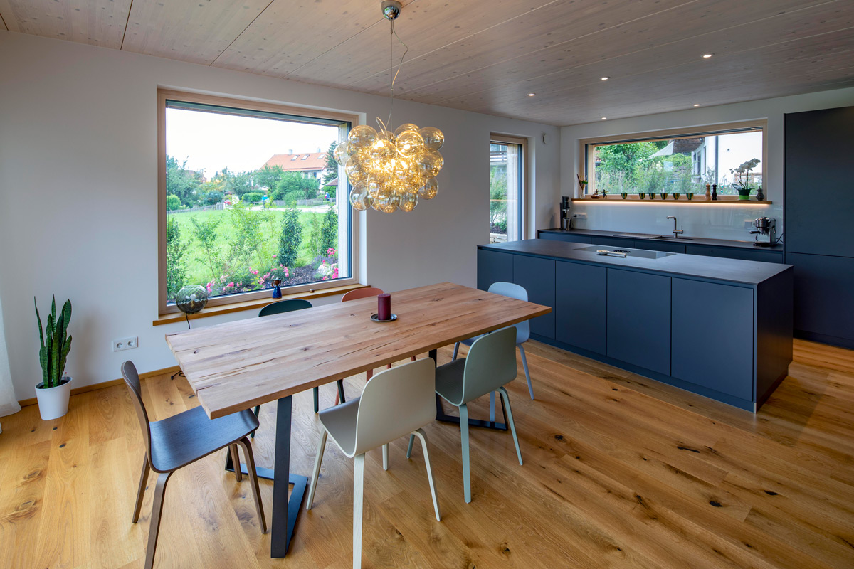 Ein modernes Esszimmer im Holzhaus in Chieming zeigt einen Holztisch mit stilvoller Beleuchtung und Blick ins Grüne.