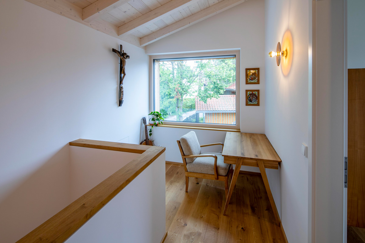 Ein heller Flur im Holzhaus Chieming zeigt einen Holztisch und Stuhl vor einem großen Fenster mit Blick auf grüne Bäume.