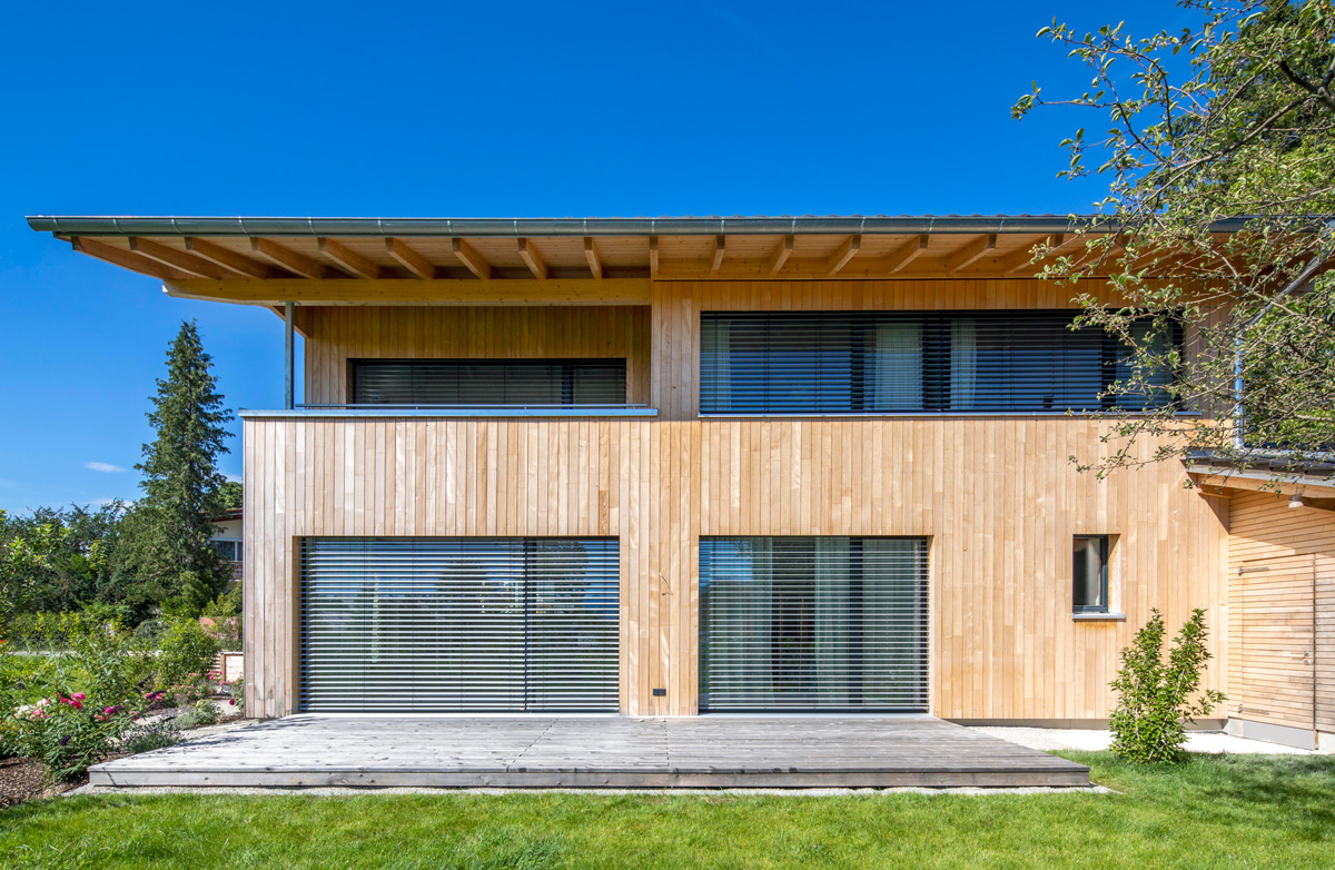 Ein modernes Holzhaus in Chieming mit großer Terrasse steht unter klarem blauem Himmel.
