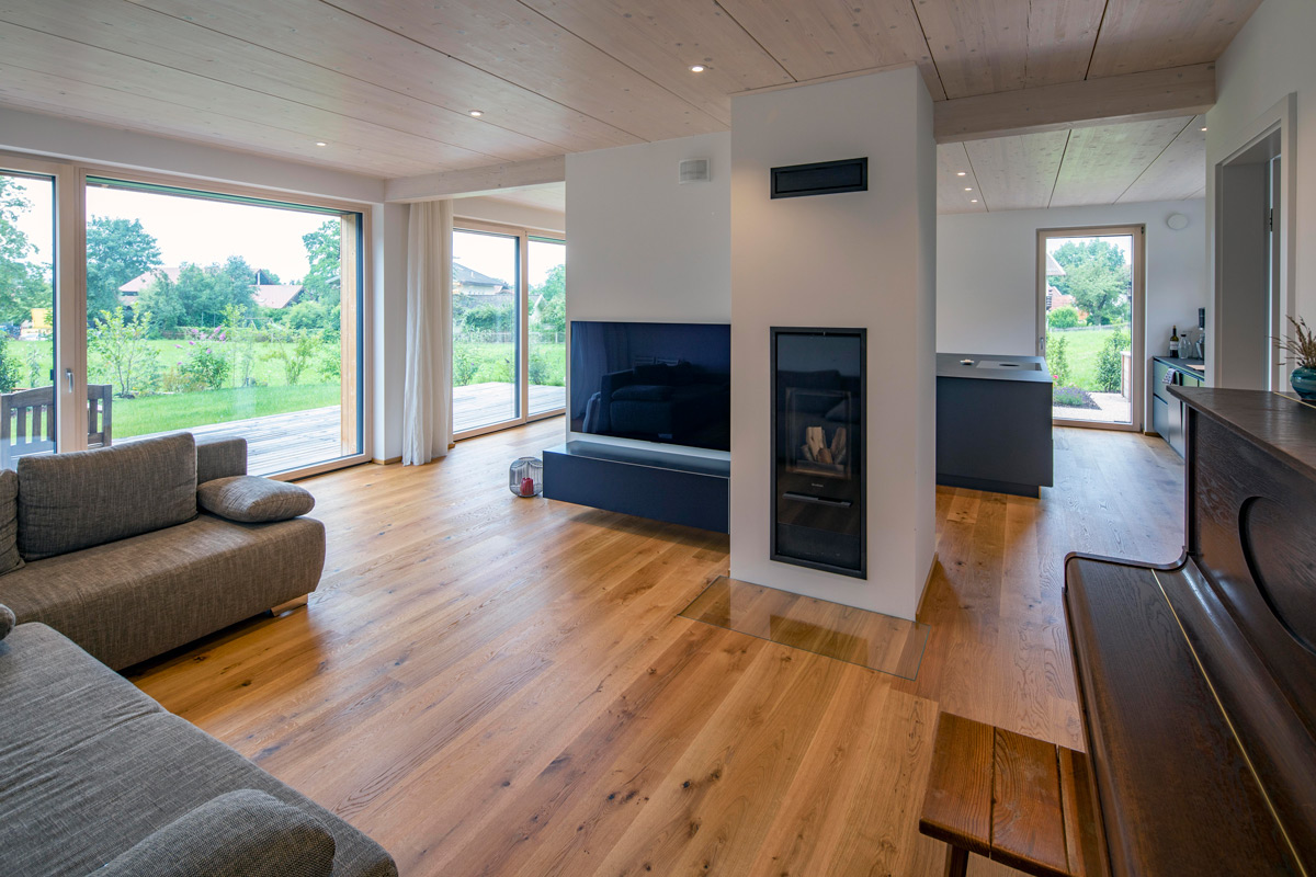 Ein modernes Wohnzimmer in einem Holzhaus in Chieming mit Holzboden, Kamin und großen Glasfenstern mit Blick ins Grüne.