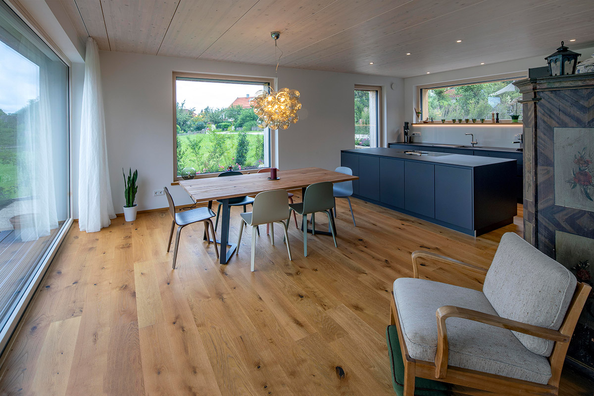 Ein modernes Holzhaus in Chieming bietet einen offenen Wohnraum mit Esstisch, Kücheninsel und großen Fenstern zum Garten.