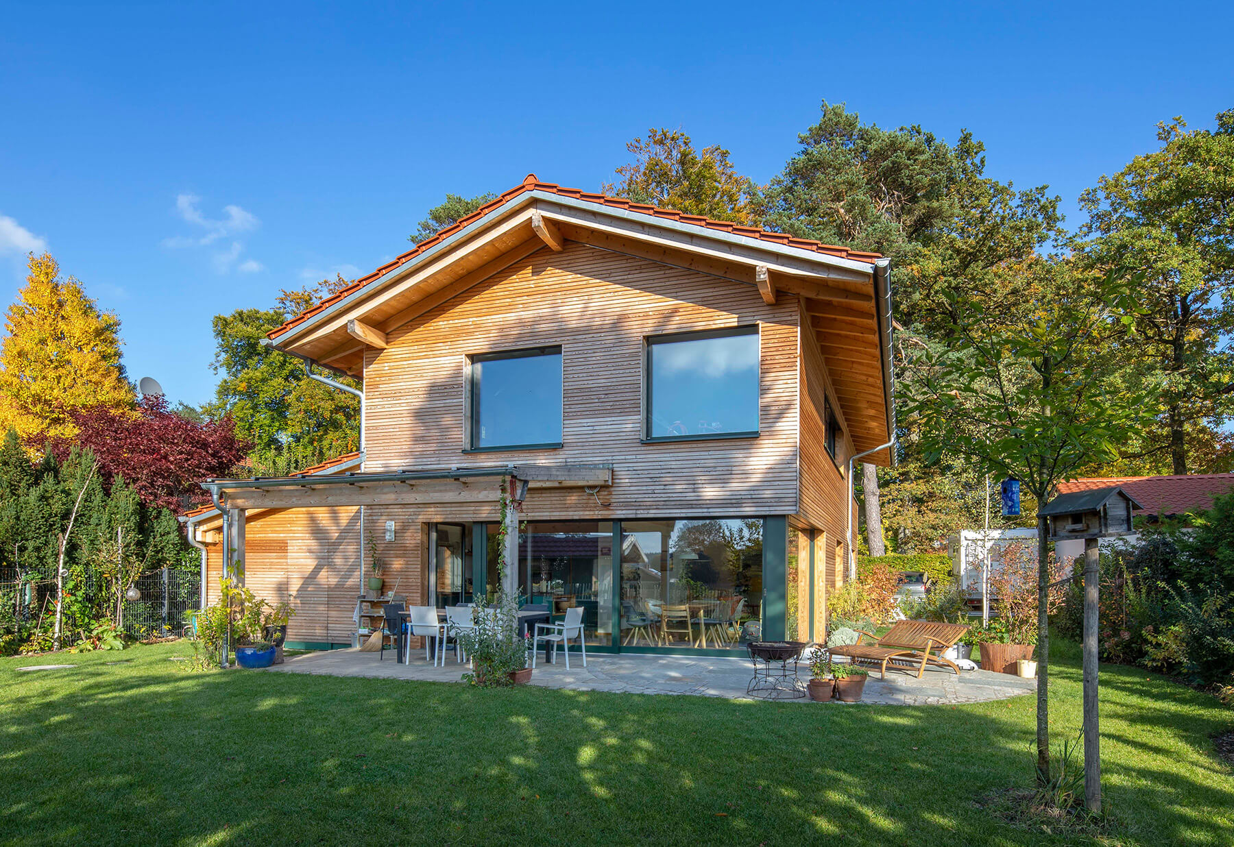 Ein modernes Holzhaus in Oberbiberg strahlt im Sonnenschein, umgeben von einem grünen Garten und herbstlichen Bäumen.
