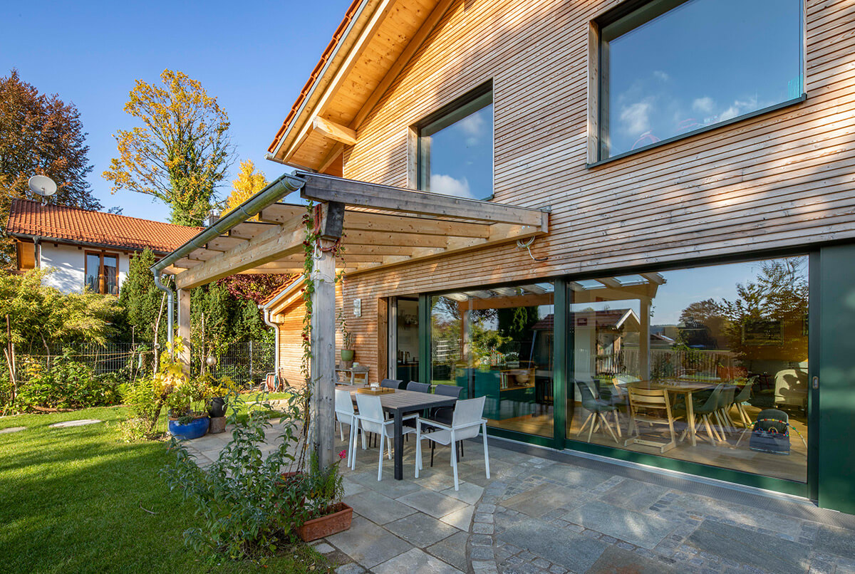 Ein modernes Holzhaus in Oberbiberg zeigt seine große Glasfront und gemütliche Terrasse bei sonnigem Wetter.