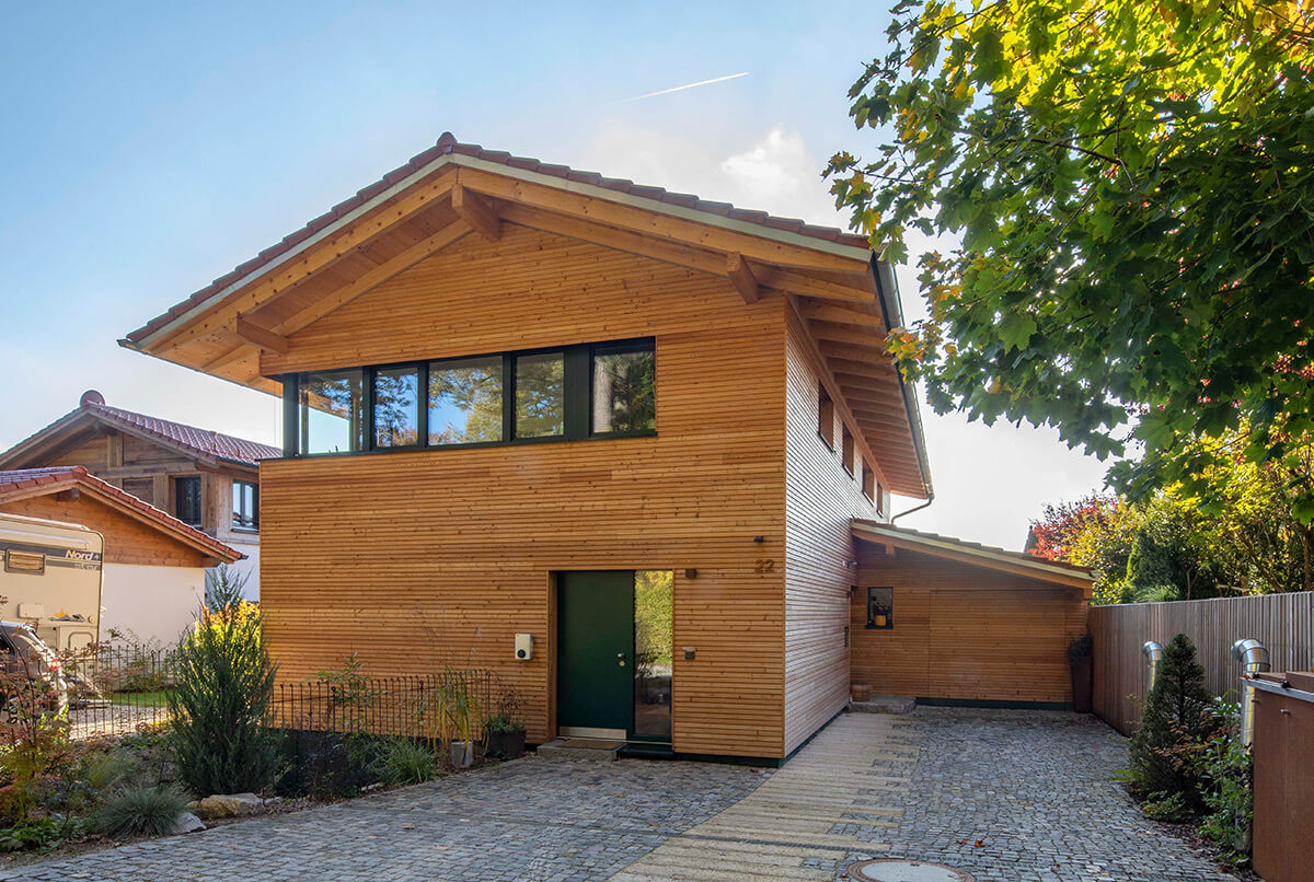 Ein modernes Holzhaus in Oberbiberg steht unter blauem Himmel, umgeben von Bäumen und einem gepflasterten Weg.