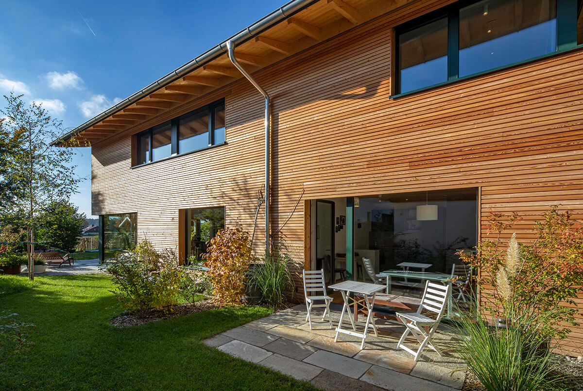 Ein modernes Holzhaus in Oberbiberg mit großer Fensterfront und gemütlicher Terrasse strahlt im sonnigen Wetter.