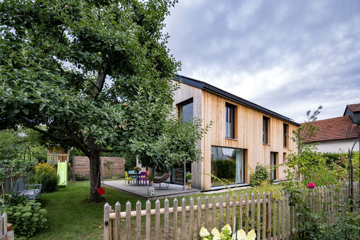 Gartenansicht mit Baum im Vordergrund, Holzfassade, große Hebeschiebetüren, Holzhaus mit Passivhauskomponenten