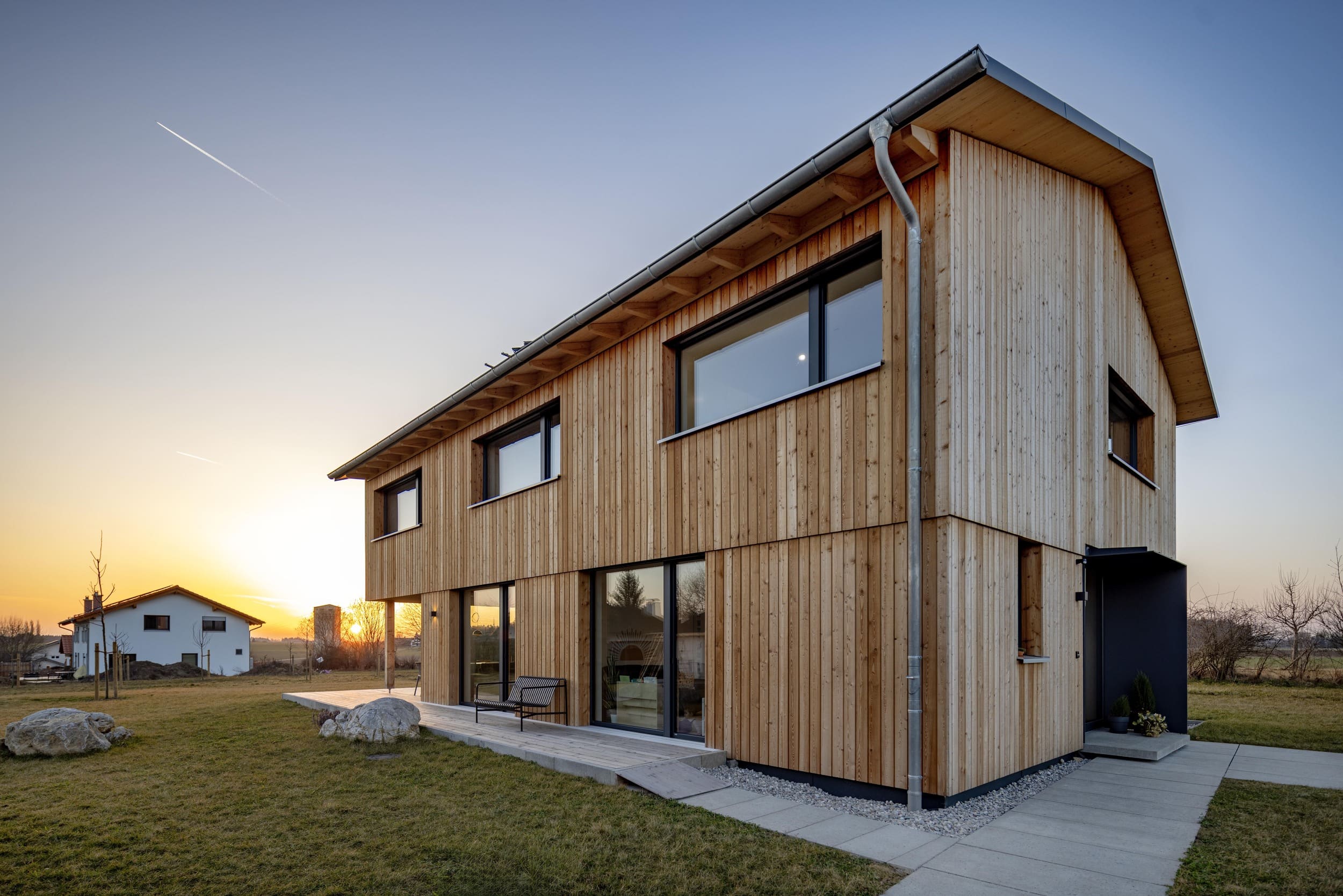Ein modernes Holzhaus steht bei Sonnenuntergang in einer ruhigen ländlichen Umgebung auf einem gepflegten Rasen.