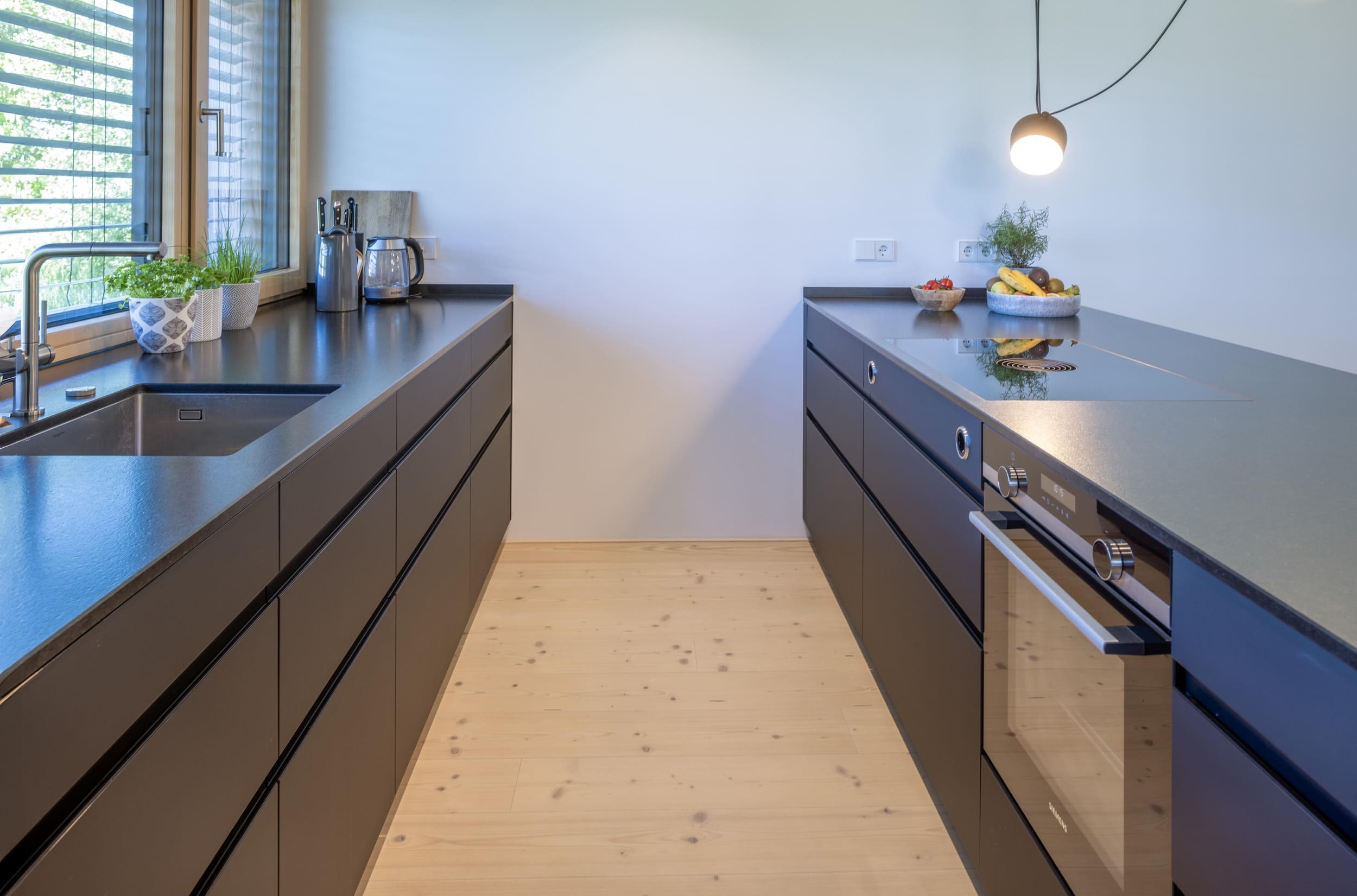 Moderne minimalistische Küche mit schwarzen Schränken, Kochfeld und frischen Kräutern auf der Fensterbank.