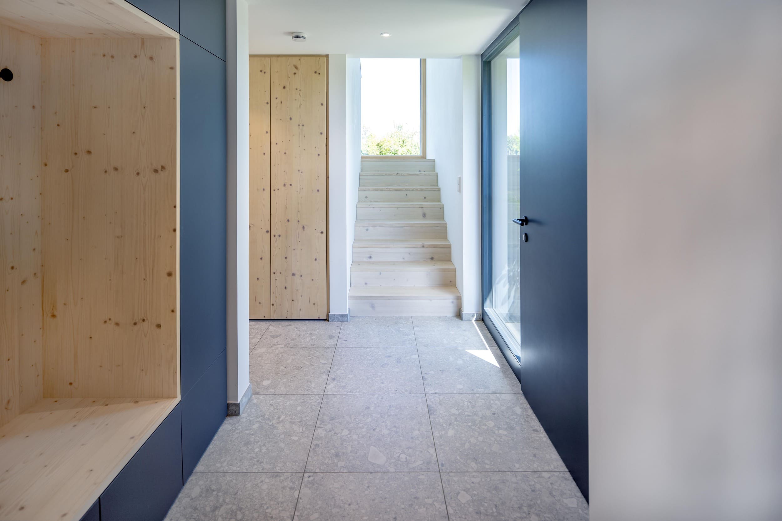 Ein moderner Flur mit hellen Fliesen und Holzverkleidung führt zu einer von natürlichem Licht erhellten Treppe.